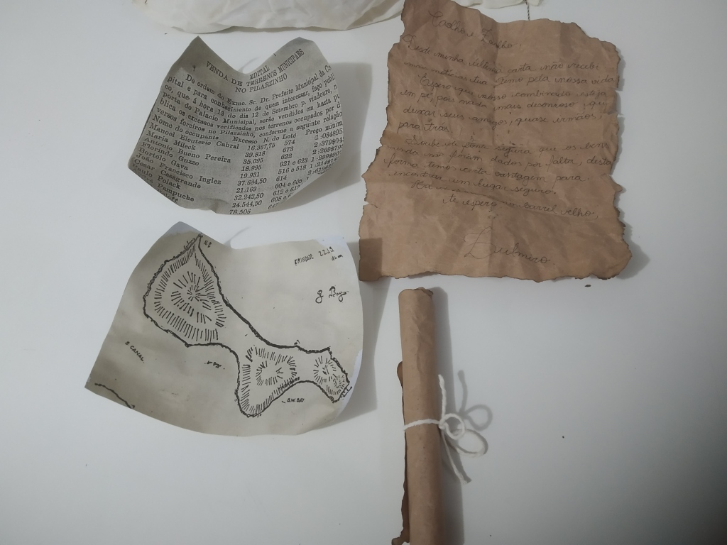 Descrição de imagem: Fotografia colorida com três documentos envelhecidos, espalhados em uma mesa de cor branca. Na parte inferior direita, documento amarrado com barbante.