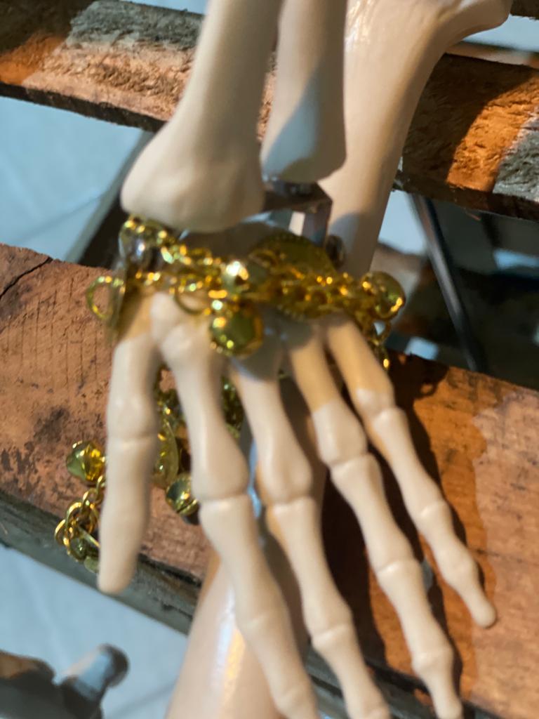 Descrição de imagem: foco em pulseira dourada entorno de uma mão esquelética, apoiada em ripas de madeira.